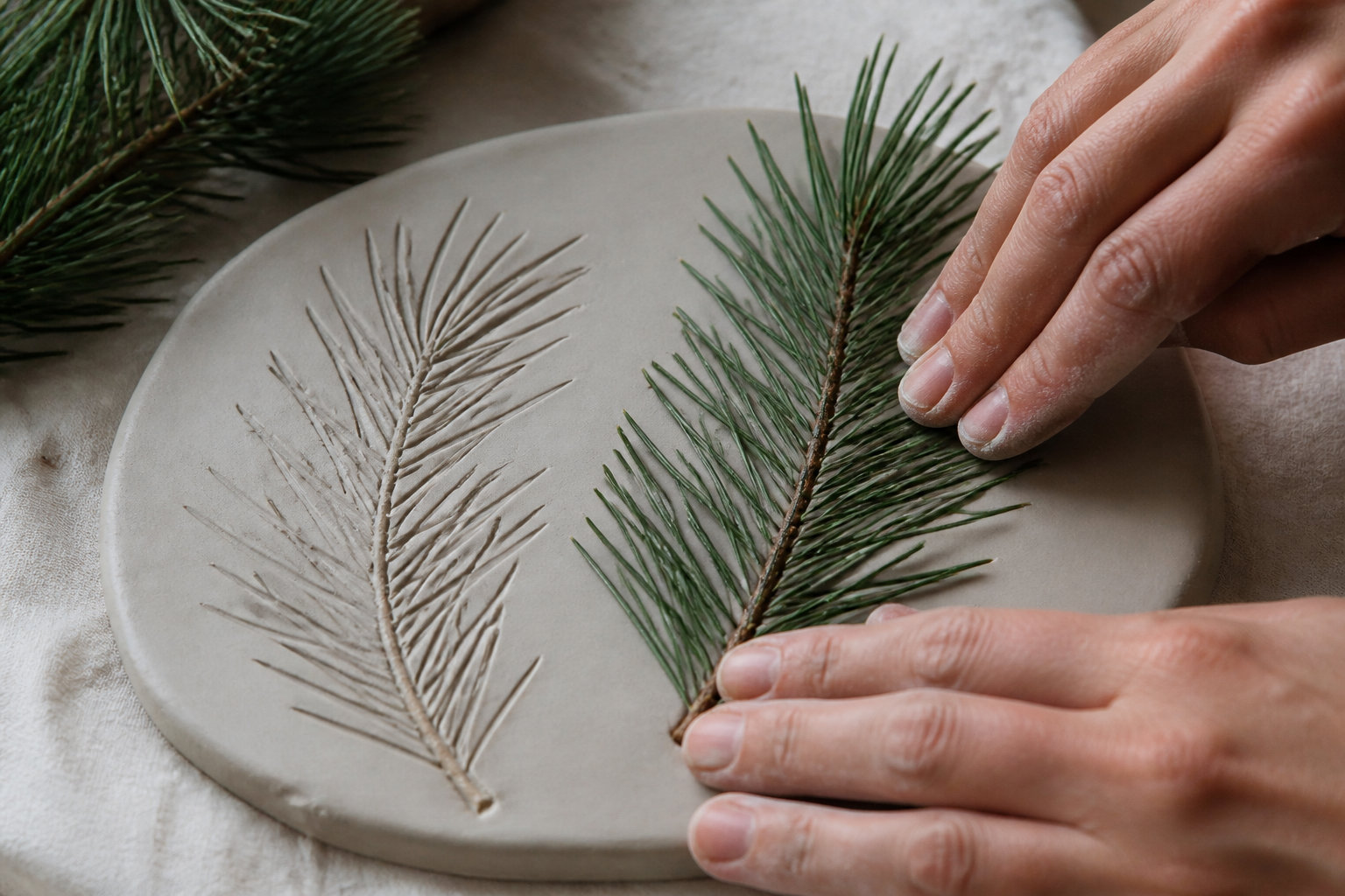 Mains imprimant une branche de sapin dans un disque d'argile encore fraîche