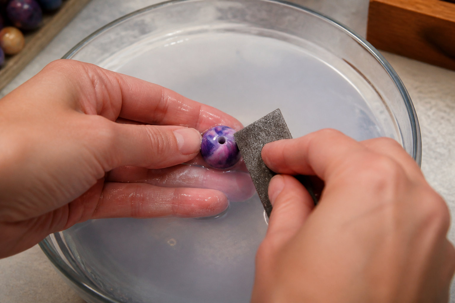 Mains ponçant à l'eau une perle violette en pâte polymère au-dessus d'un bol d'eau